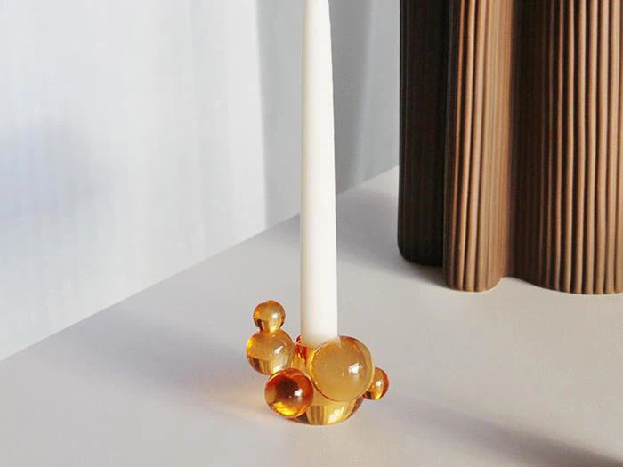 Et hvidt stearinlys i en holder med ravfarvede glaskugler, placeret på en lys flade ved siden af en brun, lodret kantet genstand.