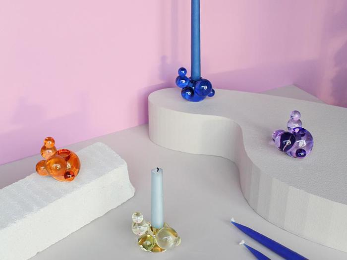 Farverige glasboble-lysestager med lys står på hvide, skulpturelle flader mod en lyserød væg. Stagerne er orange, gule, blå og lilla.