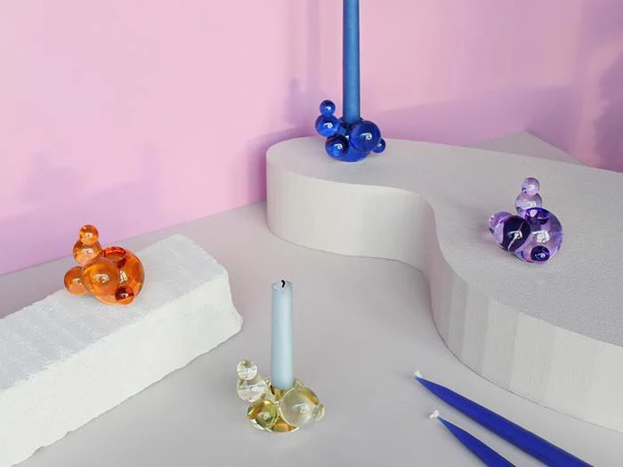 Farverige lysestager med glasbobler og blå lys arrangeret på hvide geometriske platforme med pink baggrund.