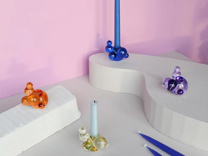 Farverige glasboble-lysestager med lys står på hvide, skulpturelle flader mod en lyserød væg. Stagerne er orange, gule, blå og lilla.