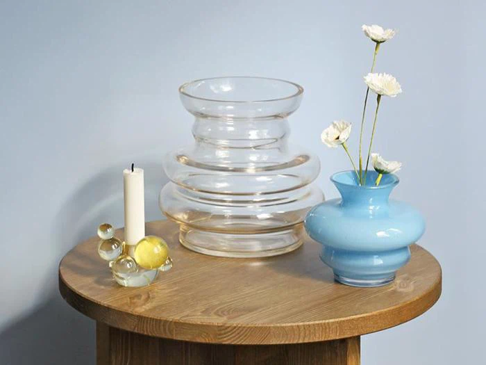 Et rundt træbord med et hvidt stearinlys, dekorative glaskugler, en klar glasvase og en blå vase med hvide blomster på en lyseblå væg.