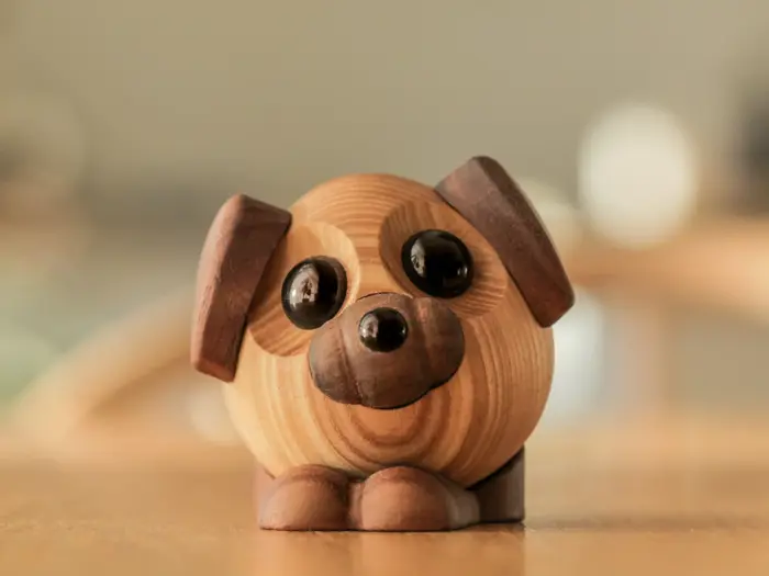 En rund hundefigur i træ med store sorte øjne, mørke ører og et smilende ansigt sidder på en lys træoverflade.
