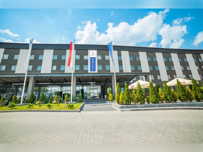 Moderne hotelbygning med flere vinduer, tre flagstænger ude foran, en anlagt indgang og udendørs siddepladser under parasoller på en solrig dag med blå himmel og skyer.