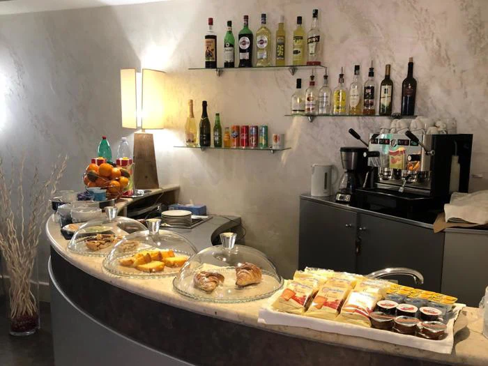 En morgenmadsbuffet med bagværk, kager, frugt og færdigpakkede snacks på en disk; flasker med alkohol og sodavand, en kaffemaskine og en frugtskål står i baggrunden.