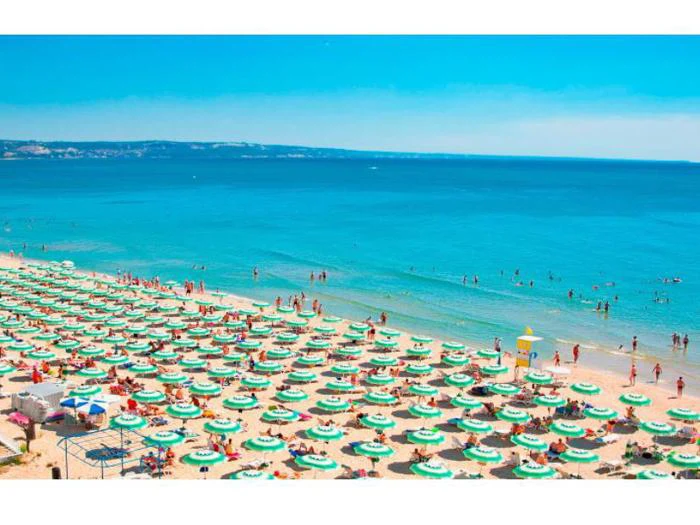 En overfyldt strand med rækker af grønne og hvide parasoller, folk, der soler sig i sandet, og mange, der bader i det klare blå hav under en solrig himmel.