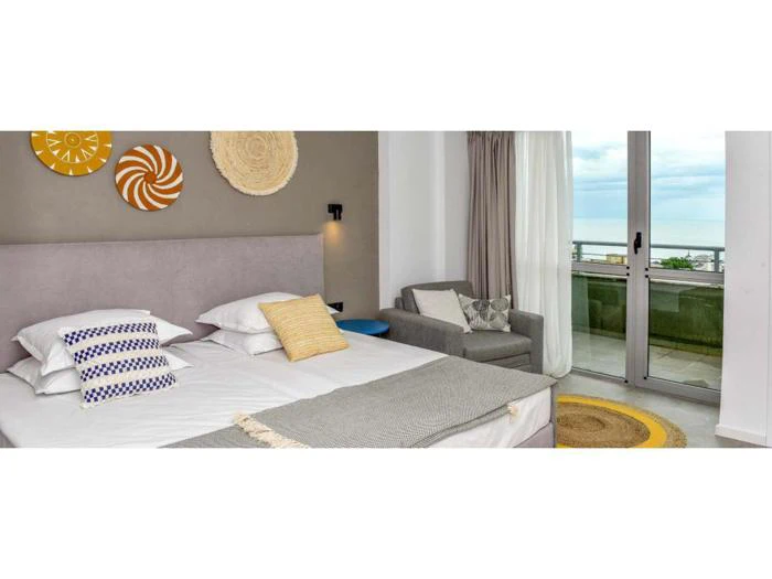 Et moderne hotelværelse med en dobbeltseng, dekorative puder, vægkunst over hovedgærdet, en lænestol og en balkon med havudsigt.