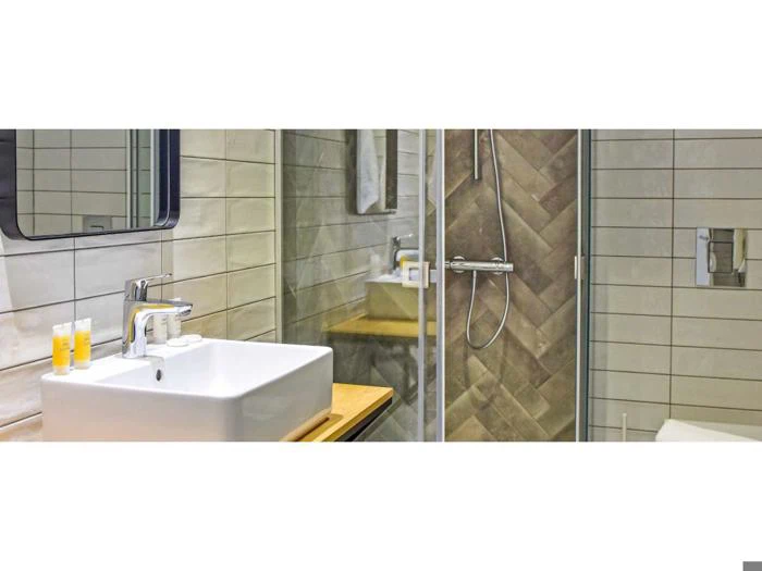 Moderne badeværelse med en firkantet hvid håndvask, vandhane, toiletartikler, et spejl og en brusekabine i glas med regnbruser og flisebelagte vægge.