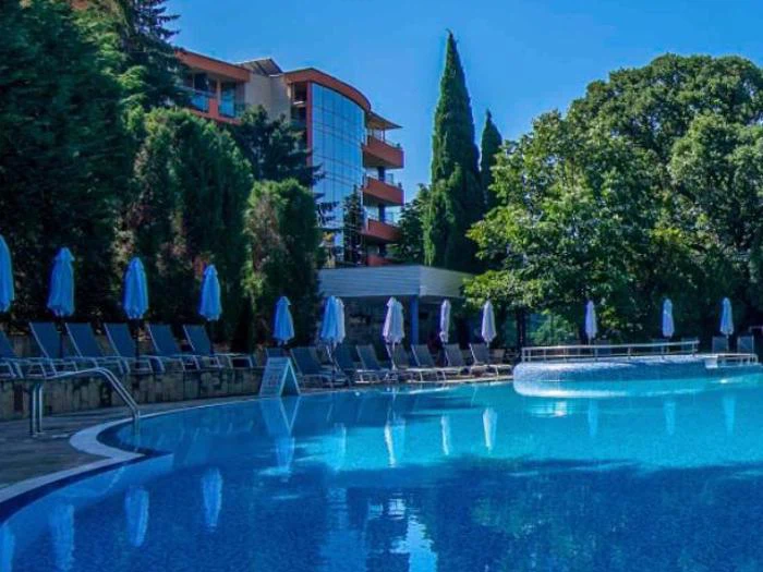 En klar blå swimmingpool omgivet af liggestole med lukkede parasoller, ved siden af et moderne hotel og frodige grønne træer under en lys himmel.