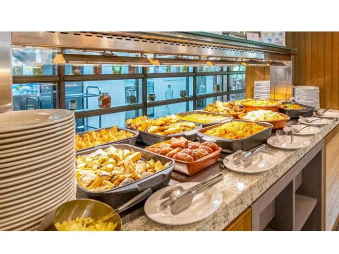 En buffetdisk med forskellige retter i bakker, bl.a. stegt mad, pasta og grøntsager, ved siden af stabler af hvide tallerkener og serveringsredskaber.