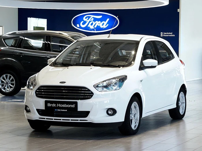 En hvid Ford hatchback er udstillet i et showroom med et stort blåt Ford-logo på væggen i baggrunden.