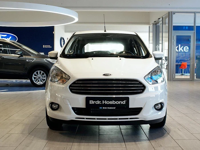 En hvid Ford er udstillet indendørs i en bilforhandlers showroom med front mod en anden mørk bil i baggrunden.
