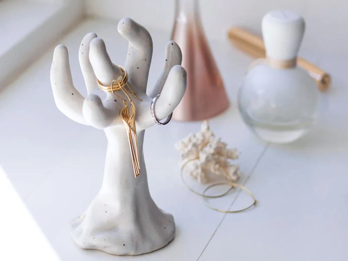 En keramisk håndformet smykkeholder viser ringe og halskæder. I baggrunden ses en lyserød vase, en hvid flaske og et stykke koral på en hvid flade.