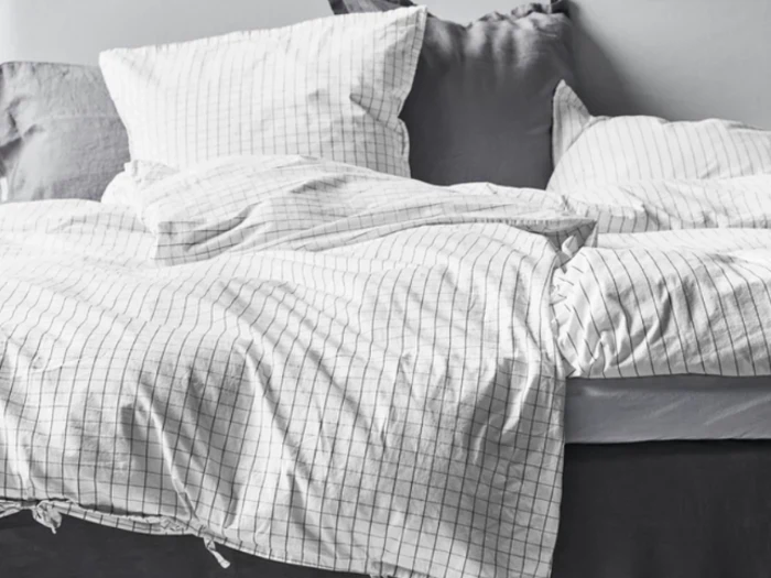 Uredt seng med hvid- og gråternet sengetøj, puder og et gråt hovedgærde mod en lys væg.