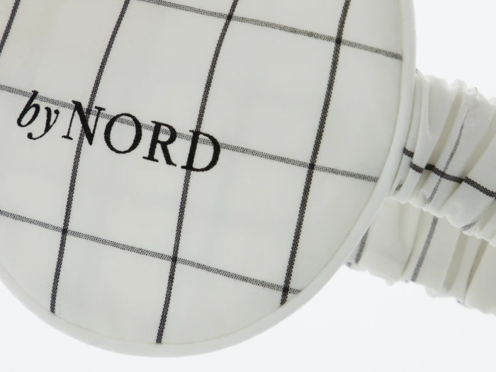 Nærbillede af en øjenmaske af hvidt stof med et sort gittermønster og ordene by NORD broderet på.