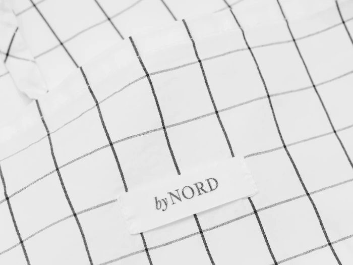Nærbillede af et hvidt stof med et sort gittermønster og en hvid label, hvor der står by NORD, syet på.