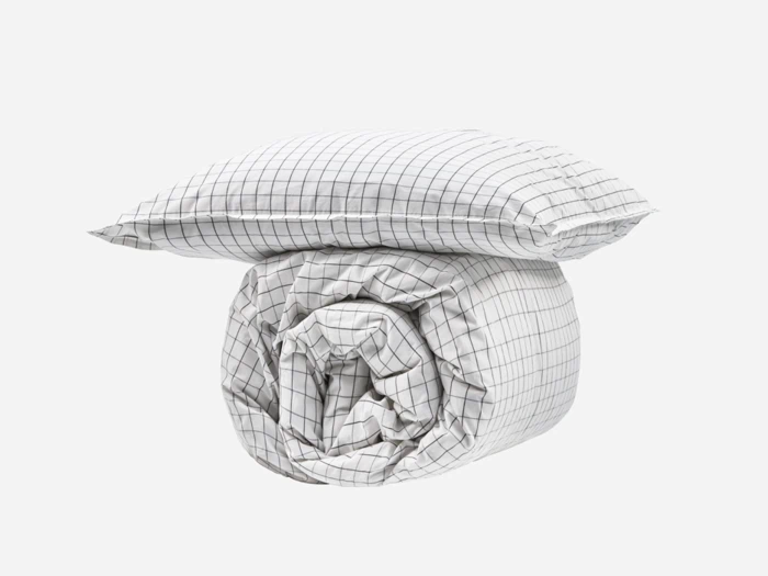 En sammenrullet dyne og en pude, begge dækket af hvidt stof med et sort gittermønster, placeret på en almindelig hvid baggrund.