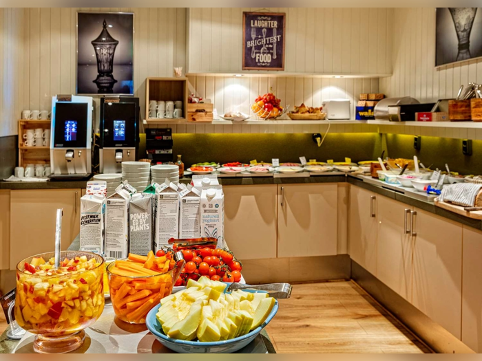 En morgenmadsbuffet med frugt, cherrytomater, gulerodsstave, mælkekartoner, morgenmadsprodukter, brød og kaffemaskiner på en bordplade i et godt oplyst køkken.