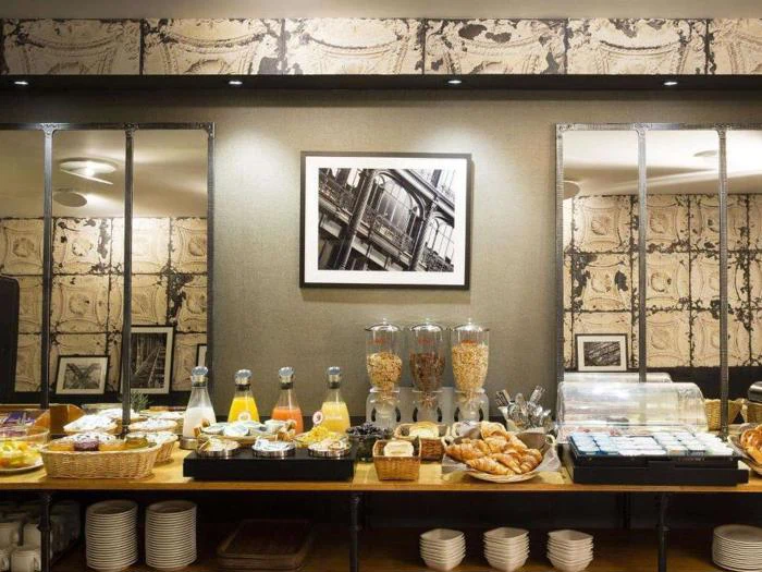 En morgenmadsbuffet med morgenmadsprodukter, kager, frugt, juice og mælk på et træbord mod en væg med to spejle og et sort-hvidt indrammet foto over.