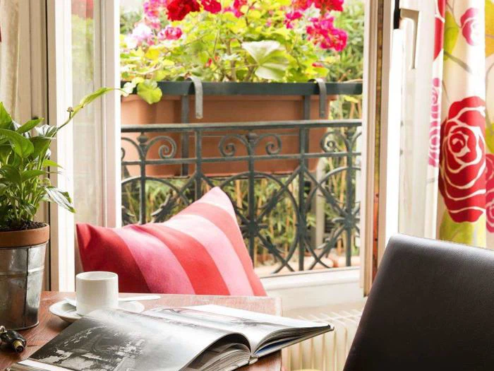 Et hyggeligt værelse med en stribet pude, en potteplante, en åben bog og en kop kaffe ved et vindue med udsigt til knaldrøde blomster på en balkon.