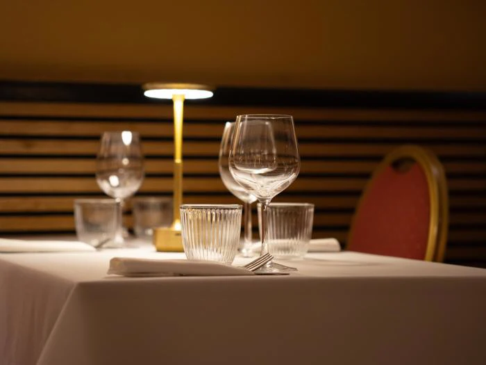 Elegant restaurantbord dækket med vinglas, vandglas, servietter og en lille lampe, med en rød polstret stol i baggrunden.