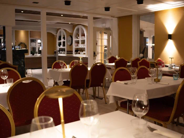 Elegant restaurantinteriør med røde og gyldne stole, hvide duge, tomme borde, vinglas og dæmpet belysning, som skaber en hyggelig og indbydende atmosfære.