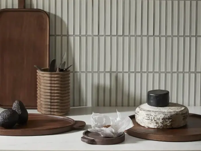 Et køkkenbord med træplader, en kurv med redskaber, avocadoer, indpakket ost og et rundt ostehjul udstillet mod en flisebelagt bagvæg.