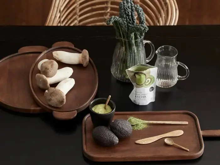 Et træbord med østershatte, avocadoer, matchapulver, matchadrik, træredskaber og glaskander med vand og bladgrønt.