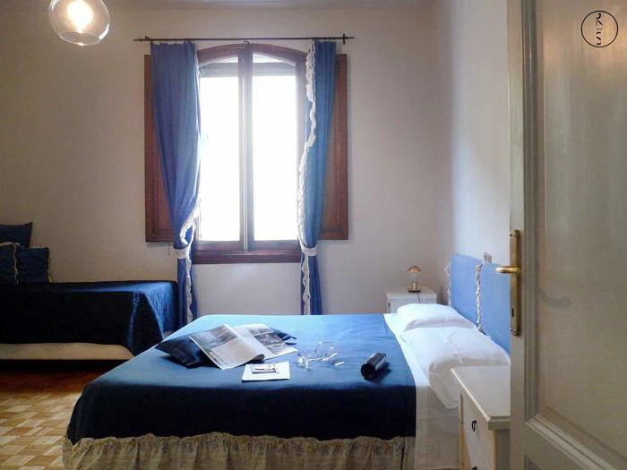 Lyst soveværelse med et stort vindue, blå gardiner, en dobbeltseng med blåt sengetøj, et natbord og et magasin og briller placeret på sengen.