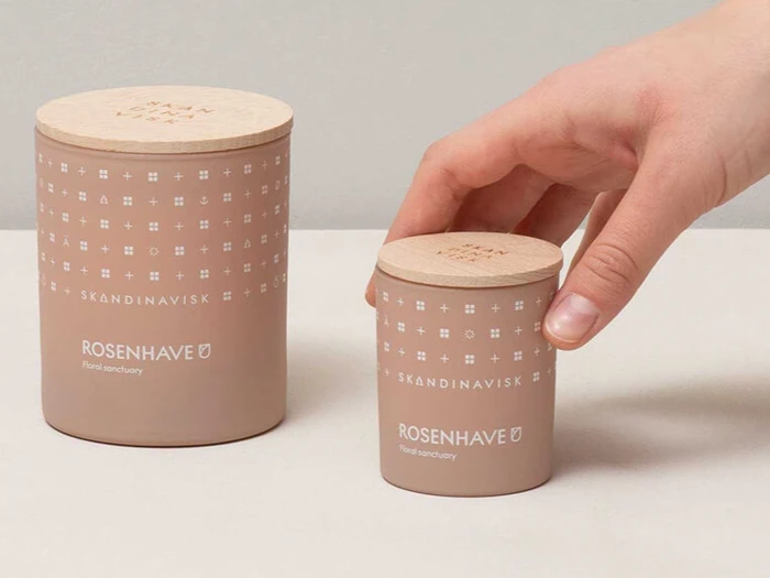 En hånd rækker ud efter et lille, lyserødt SKANDINAVISK ROSENHAVE-lys med trælåg ved siden af et større, matchende lys på en lys flade.