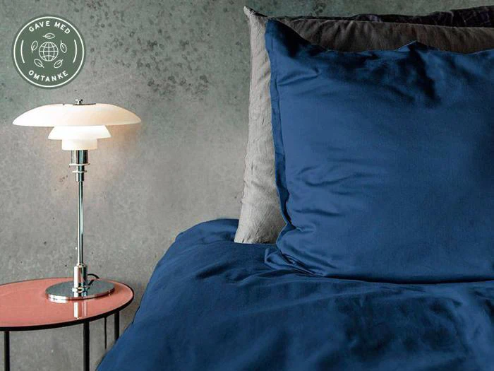 En pænt redt seng med mørkeblå lagner og grå puder ved siden af et lille rundt bord med en moderne lampe mod en struktureret grå væg.