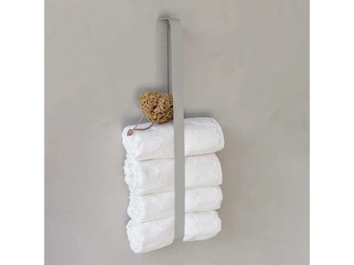 Et væghængt metalstativ holder fire rullede hvide håndklæder og en naturlig havsvamp mod en almindelig grå væg.