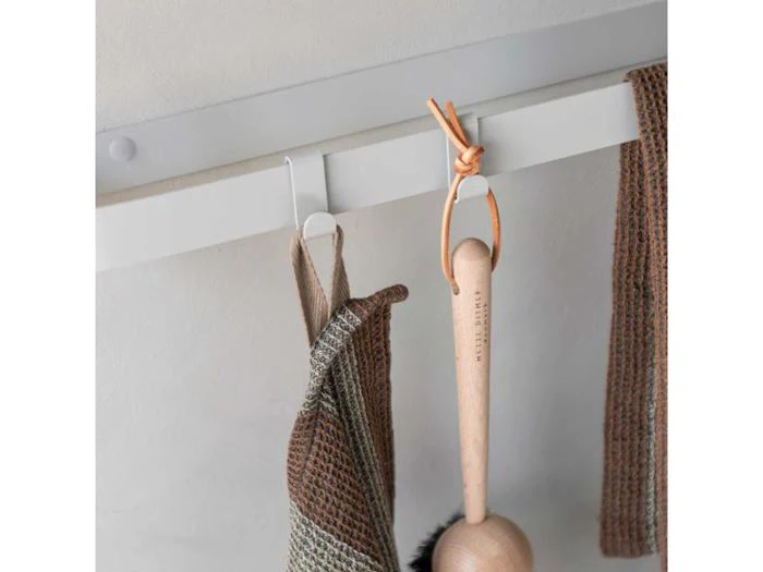 En træbørste med læderstrop og to brune viskestykker hænger på kroge på et hvidt vægstativ.