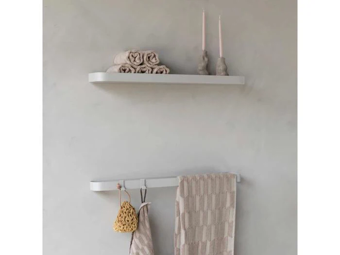 Minimalistisk badeværelsesindretning med en hvid hylde med rullede håndklæder og to stearinlys, og en håndklædeholder nedenunder med et beige håndklæde og hængende badetilbehør.