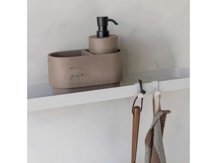 En beige sæbedispenser og en matchende beholder står på en hvid hylde med kroge under, der holder en brun strop og en netpose.