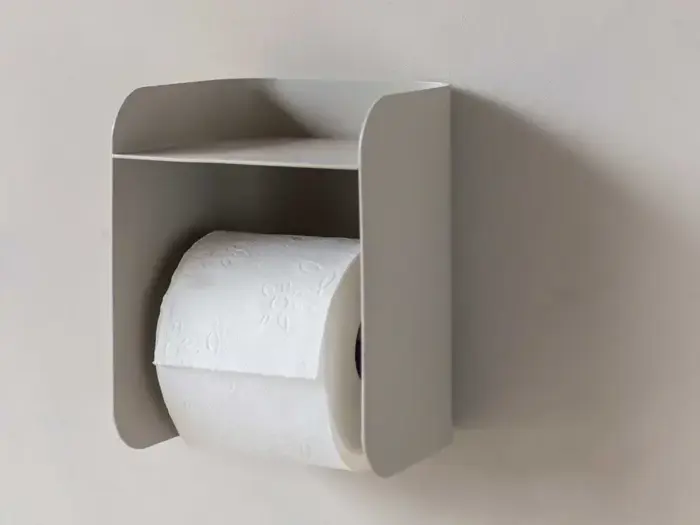 En rulle toiletpapir er placeret i en minimalistisk, vægmonteret holder med en lille hylde over, der er fastgjort til en lys væg.