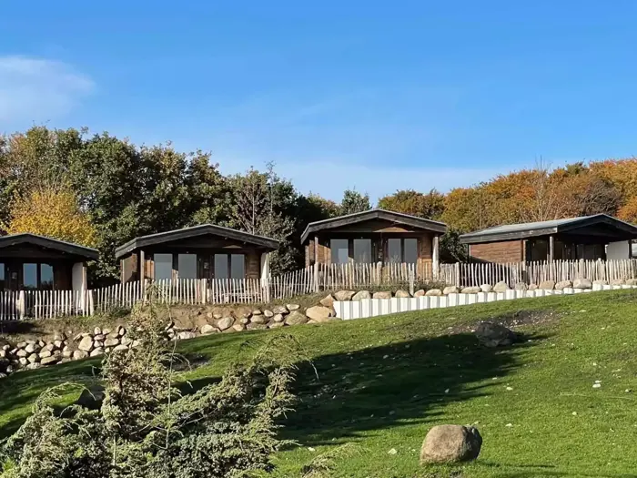 Tre træhytter med store vinduer ligger på en græsklædt bakke, omgivet af et træhegn, klipper og træer under en klar blå himmel.