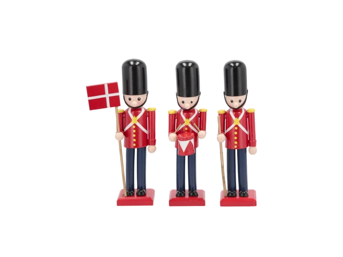 Tre legetøjssoldater af træ i røde og blå uniformer står på rad og række; en holder et dansk flag, en anden har en tromme, og den tredje holder et gevær.