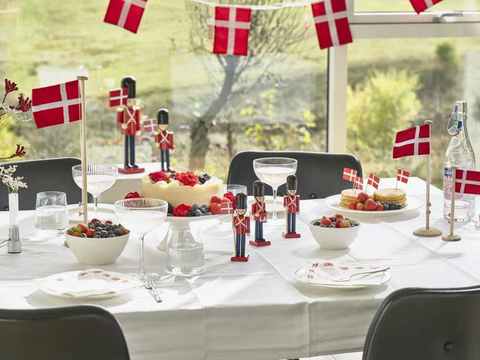 Et festligt bord pyntet med danske flag, legetøjssoldater, kager, bær, glas og tallerkener, dækket ved store vinduer med udsigt til haven udenfor.