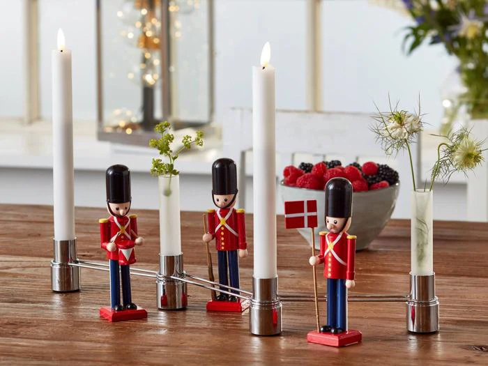 Fire legetøjssoldater holder høje hvide lys og et dansk flag på et træbord med en skål med bær og nogle små blomsterarrangementer i baggrunden.