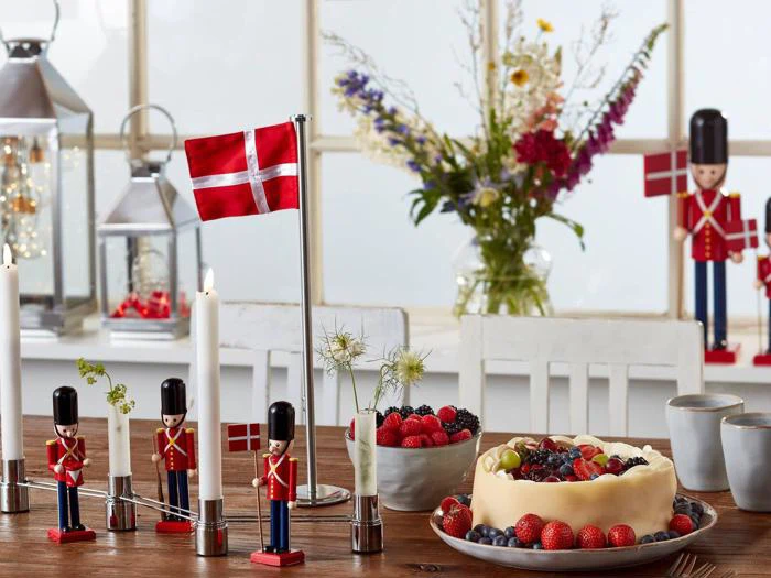 Et bord dækket med et dansk flag, legetøjssoldater, stearinlys, friske bær, en kage med frugt på toppen og en vase med blomster i baggrunden. Lanterner og små danske flag giver et festligt præg.