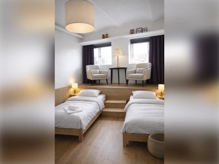 Et hotelværelse med to enkeltsenge på et lavere niveau og to lænestole på en forhøjet træplatform.