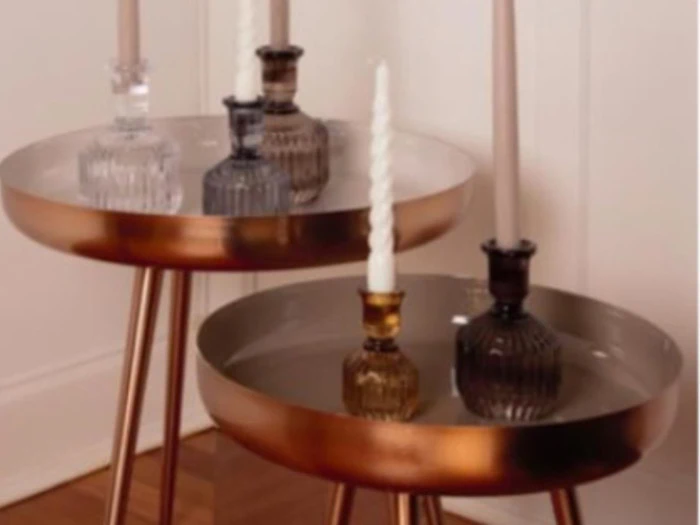 To runde, gyldne sideborde med metalben holder glaslysestager og snoede lys, opstillet mod en hvid væg på et trægulv.