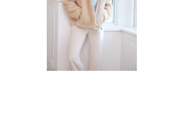 En person står indendørs ved et vindue iført en beigefarvet jakke og hvide bukser. Det øverste af ansigtet er uden for billedet.