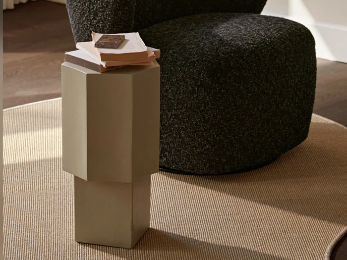 Et lille, geometrisk sidebord med et par bøger på står på et beige tæppe ved siden af en mørk stol med struktur i et blødt oplyst rum.