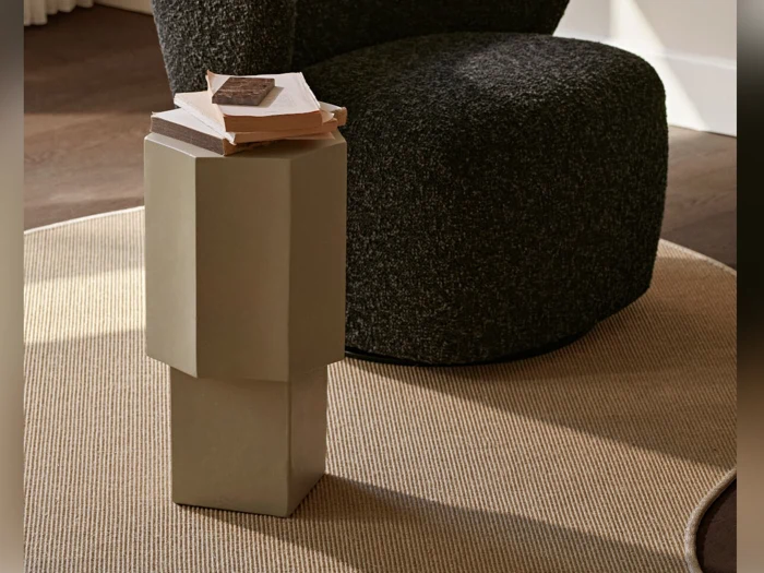 Et lille, moderne beige sidebord med bøger på står på et lysebrunt tæppe ved siden af en mørk stol med struktur i et blødt oplyst rum.