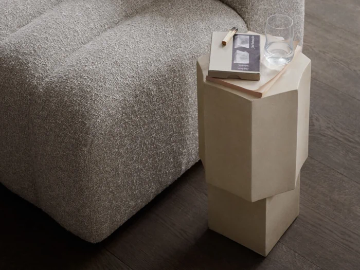 En grå sofa med struktur ved siden af et moderne beige sidebord, hvor der står en bog, en blyant og et glas vand på et mørkt trægulv.