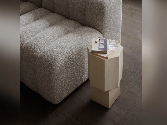 En grå sofa med struktur står ved siden af et beige, geometrisk sidebord, hvorpå der ligger en lukket bog, et par briller og et glas vand på et mørkt trægulv.