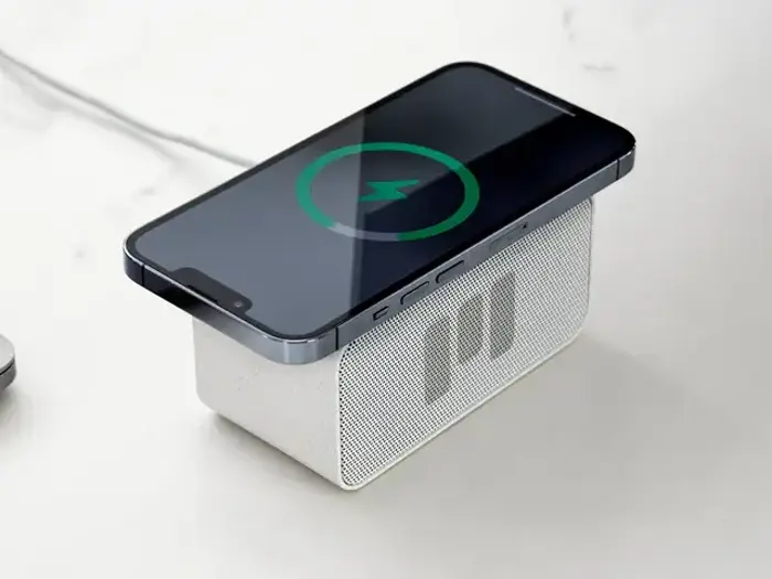 En smartphone oplades trådløst oven på en lille hvid rektangulær højttaler, der er placeret på en lys overflade.