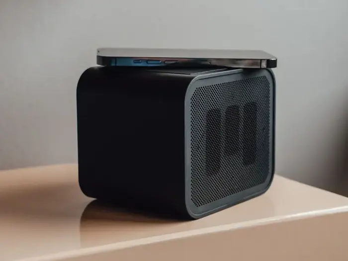 En smartphone hviler oven på en moderne, rektangulær sort trådløs højttaler med et perforeret frontgitter, der er placeret på en lys overflade.