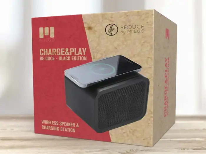 En æske med MIIEGO Charge&Play Re:duce Black Edition, en trådløs højttaler og ladestation, udstillet på en lys træoverflade.
